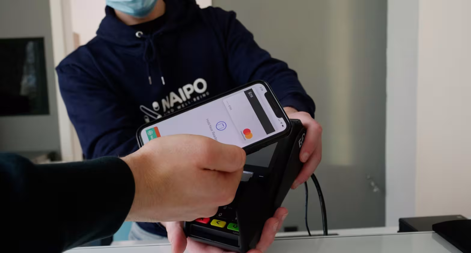 Person mit Maske hält Kartenleser, Hand bezahlt kontaktlos am Terminal