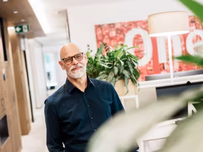 Foto: Älterer Mann mit Brille im Büroflur, Pflanzen und rotes Plakat im Hintergrund.