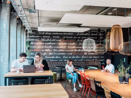 Helles Café mit großen Fenstern; mehrere Personen arbeiten an langen Holztischen.